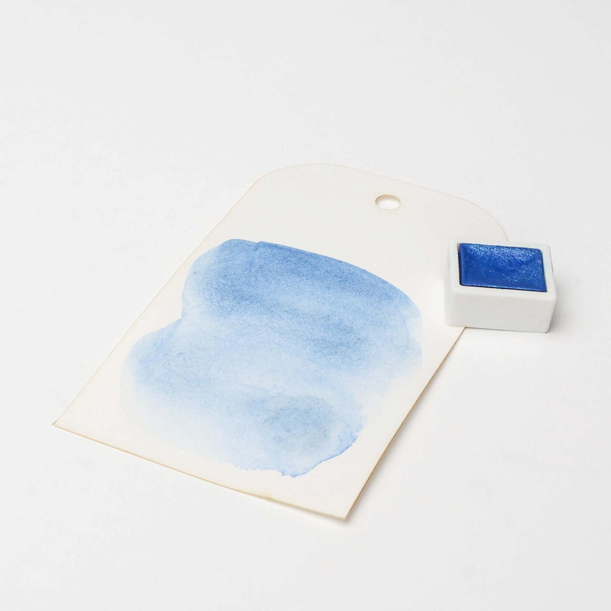 Ein blau aquarelliertes Farbmuster mit satten Farbpigmenten von Rico Design steht neben einem kleinen blauen Aquarelltopf auf weißem Hintergrund.