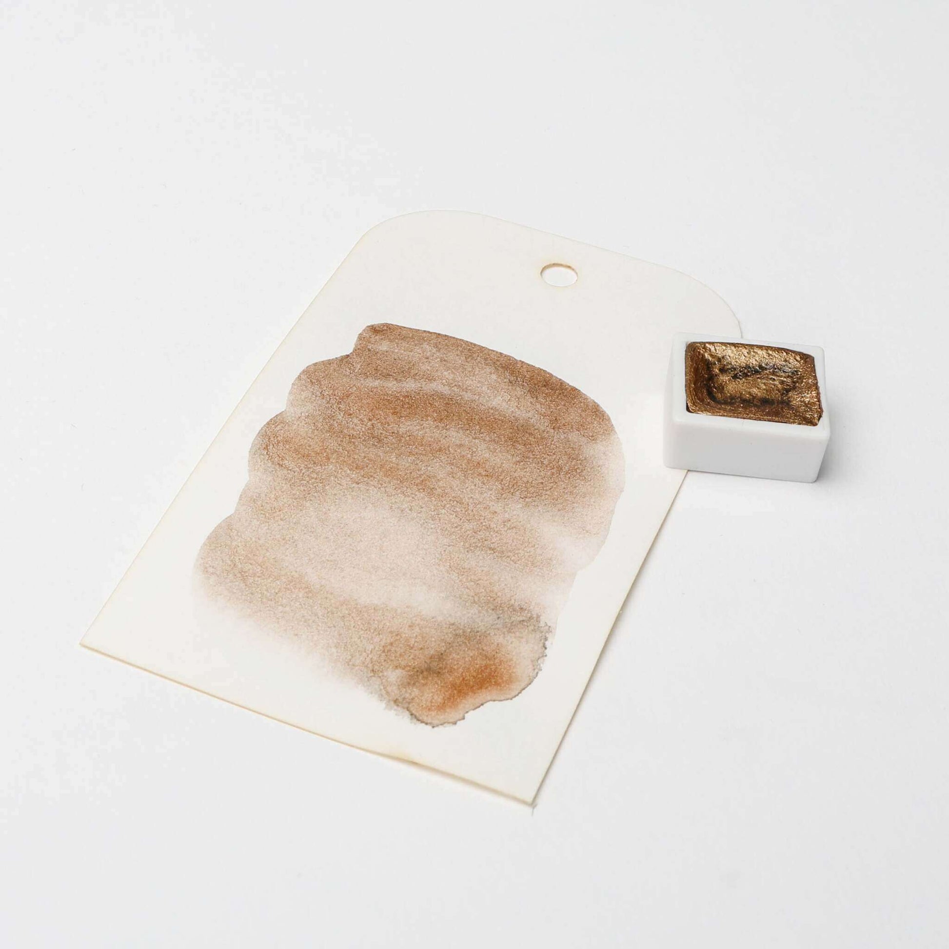 Kleine weiße Pfanne mit braunem Aquarell von Rico Design mit hoch konzentrierten Farbpigmenten, abgebildet neben einem gemalten Muster auf einem weißen Anhänger mit Loch.