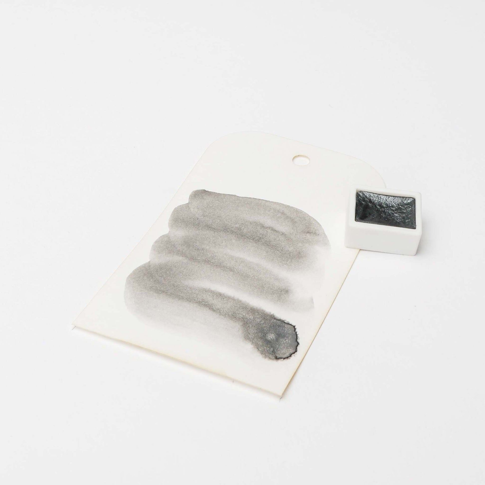 Ein schwarzer Aquarelltopf von Rico Design steht auf einem weißen Hintergrund neben einem Farbmuster mit schwarzen Pinselstrichen aus hoch konzentrierten Farbpigmenten.