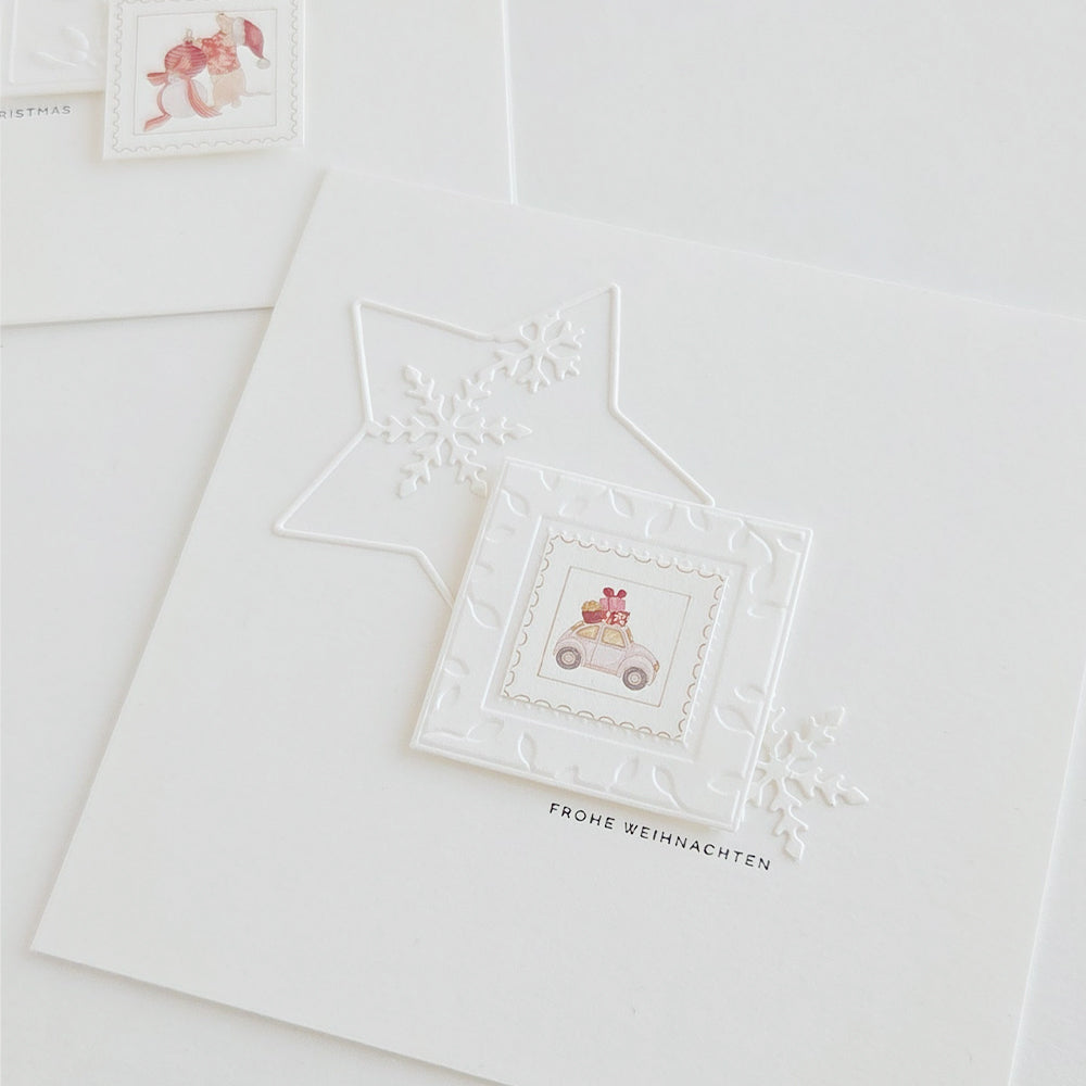 Eine quadratische, weiße Grußkarte mit einem geprägten Stern, einer Schneeflocke und einer kleinen Illustration eines Autos, das Geschenke transportiert, hergestellt mit der Prägevorlage „Cut & Emboss Folder“ von Stanzsysteme.