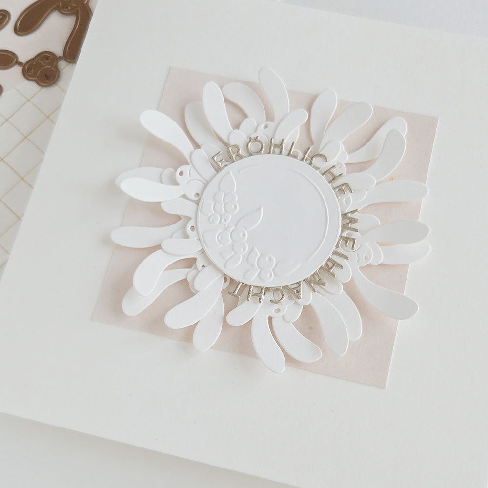 Eine Dekoration aus weißem Papier in Form einer Blume mit floralen Etiketten, auf deren mittlerem Kreis der deutsche Text „Fröhliche Weihnachten“ geprägt ist.