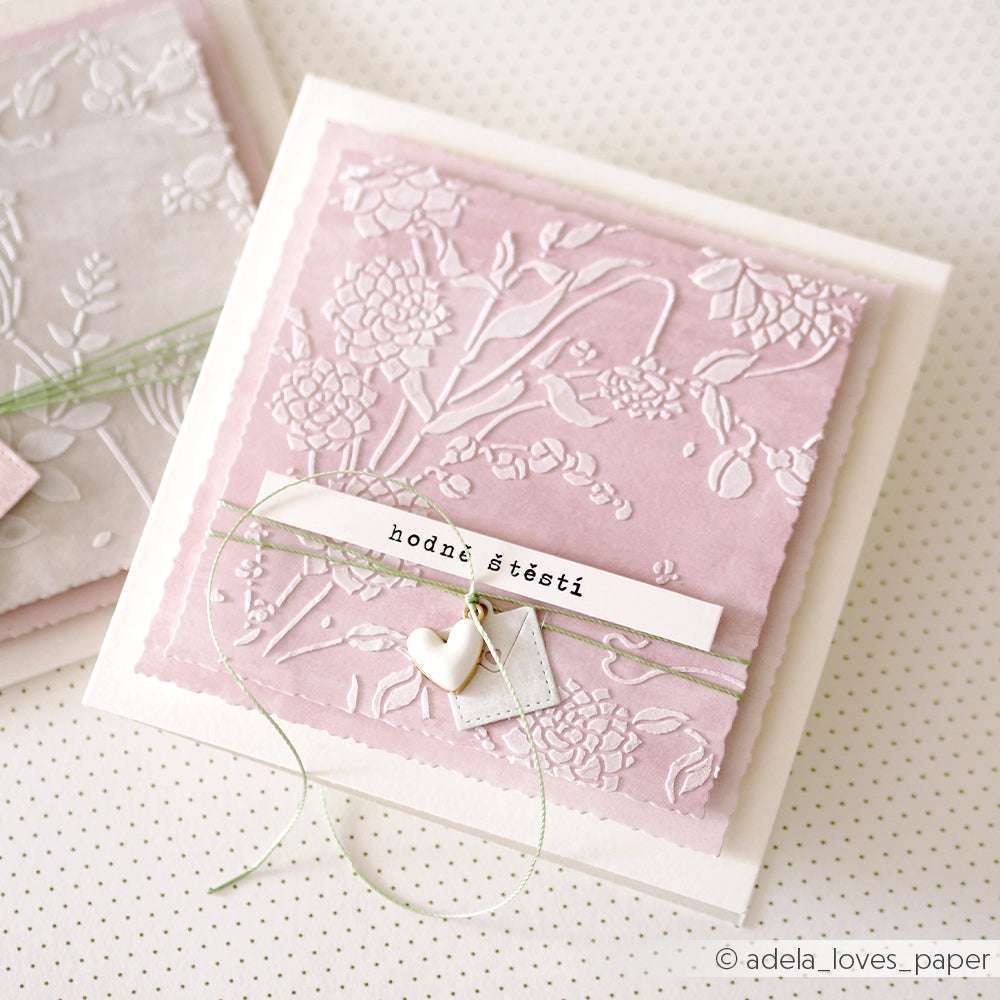 Die quadratische, pastellrosa Grußkarte von Alexandra Renke ist mit einem geprägten Blumenmuster, einer grünen Kordel und zwei weißen Herzanhängern versehen, die mit Schablonen Papierprojekten und einem Stencil Dahlienwirbel für elegante Details gefertigt wurden.