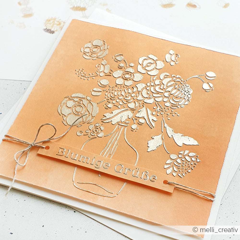 Pfirsichfarbene Karte von Alexandra Renke mit geprägten Blumen in einer Vase, die mit wiederverwendbaren Schablonen hergestellt wurden, und "Blumige Grüße" in Deutsch auf einem erhabenen Streifen.
