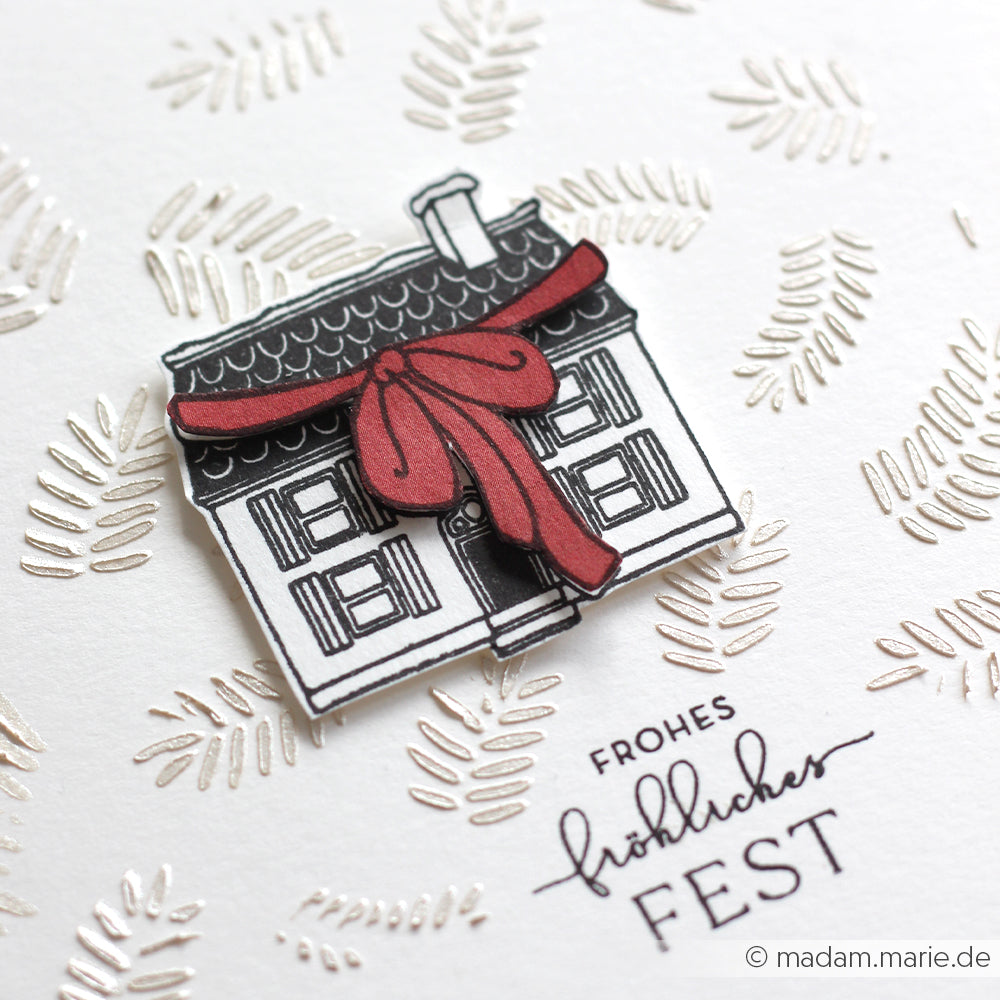 Ein Papierhaus mit einer roten Schleife sitzt auf einer weißen Karte, verziert mit goldenen Blättern, deutschem Feiertagstext und der eleganten Schablone Tannenzweige von Alexandra Renke für festliche Projekte.