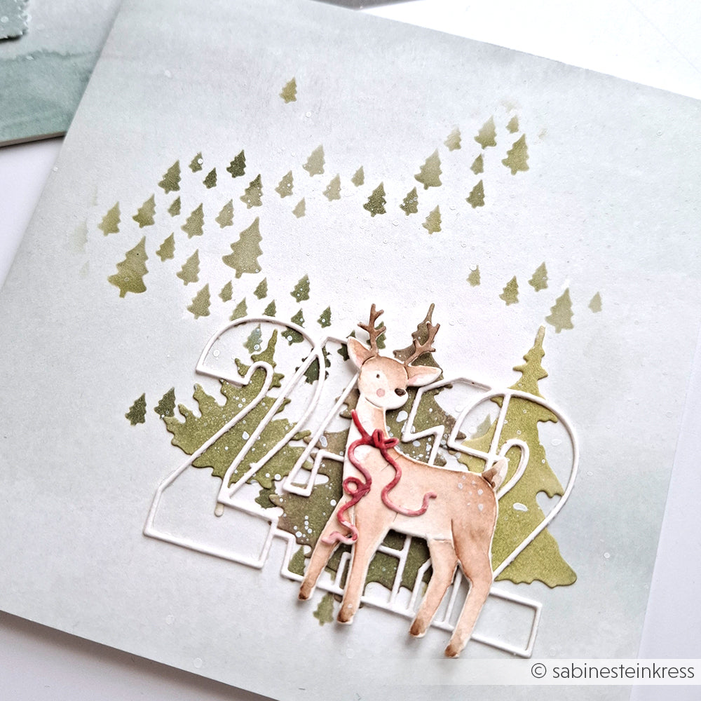 Handgefertigte Karte mit Rentier, Bäumen und "2022"-Ausschnitt auf verschneitem Hintergrund, hergestellt mit Alexandra Renke Stencil Freudige Tannenlandschaft Schablonen für einzigartigen, wiederverwendbaren Weihnachts-Charme.