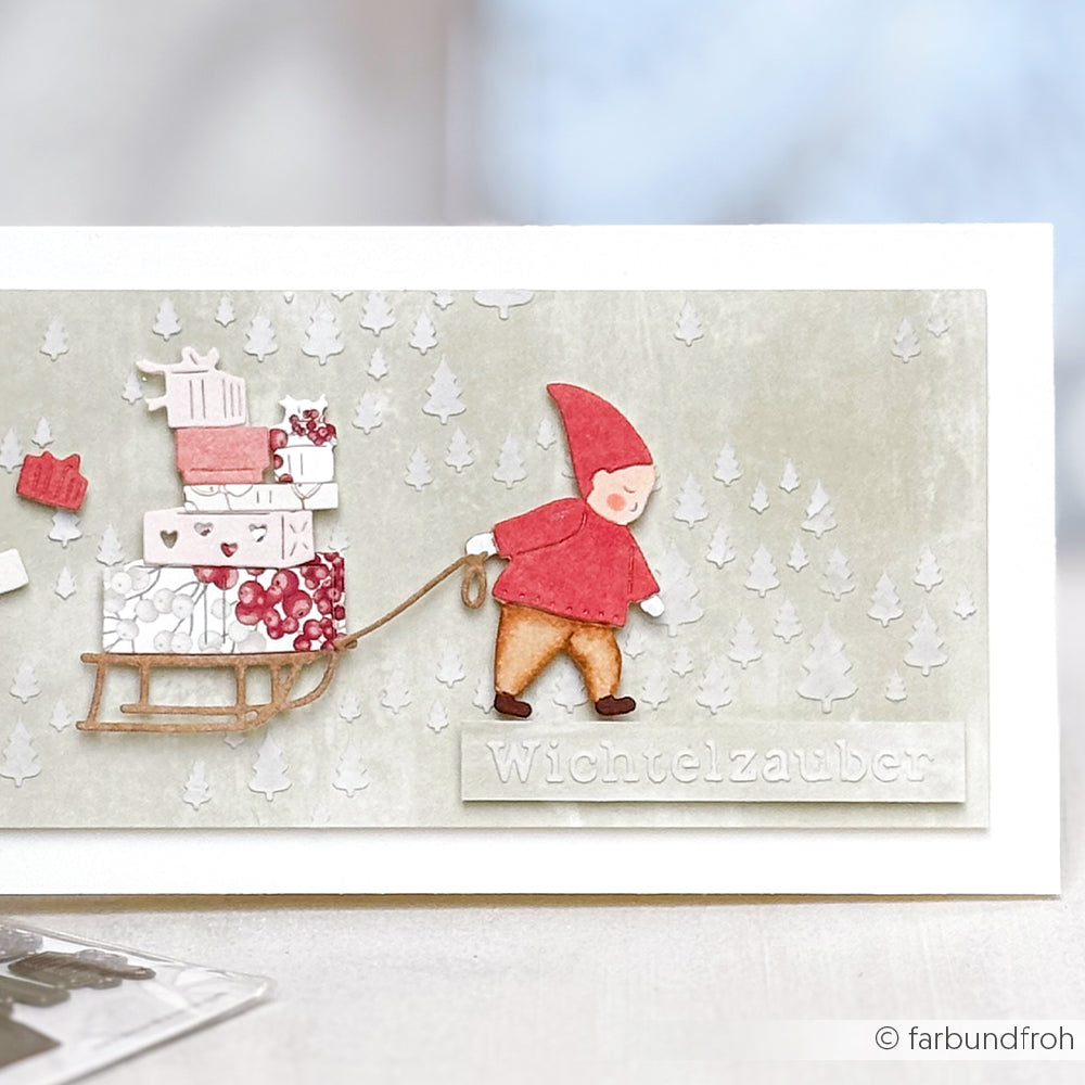 Handgefertigte Karte mit einem Gnom, der einen Geschenkeschlitten zieht, Schneeflocken und dem Text "Wichtelzauber", hergestellt mit Alexandra Renke Schablonen Papierprojekten wie der Schablone Freudige Tannenlandschaft für festlichen Charme.