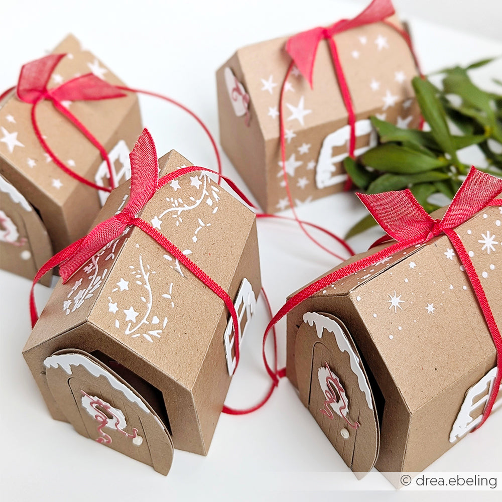 Kleine braune Geschenkschachteln, verziert mit Alexandra Renkes Schablone Ranken, roten Bändern und Grünzeug, ruhen auf einer weißen Fläche. Die hübschen weißen Motive werden mit der wiederverwendbaren Schablone Musterstreifen hergestellt und sorgen für zusätzlichen Charme.