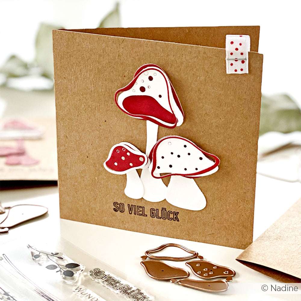 Braune Karte mit drei rot-weißen Pilzen, die mit einem Klarsichtstempelset von Alexandra Renke hergestellt wurden und unter denen der Text SO VIEL GLÜCK steht.