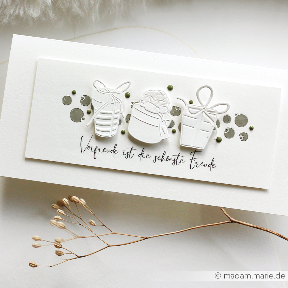 Weiße Karte von Alexandra Renke mit drei geprägten Geschenkmotiven, deutschem Text, grünen Punkten, Schablonen-Details und einer getrockneten Pflanze auf weißem Hintergrund.