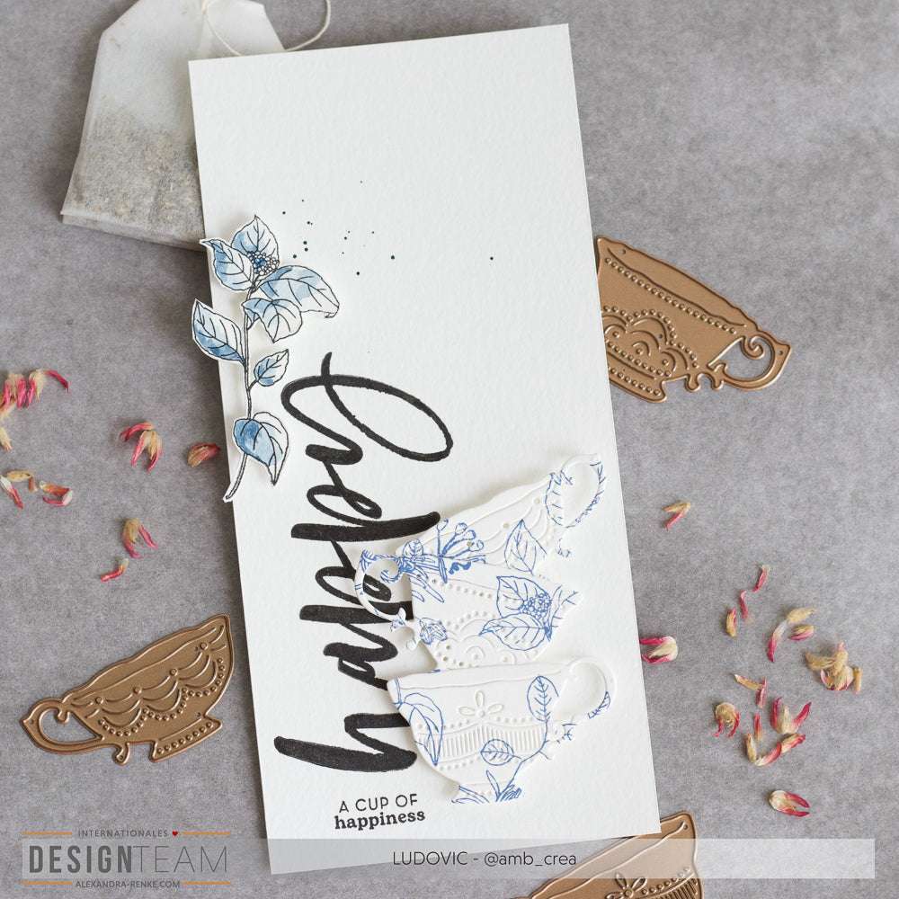 Eine handgefertigte Karte von Alexandra Renke mit blauer Teetasse, floralen Akzenten, "Glück", gestanzten Metallformen, Details aus dem Clear Stamp Set und zarten Blumenkranz-Designs.