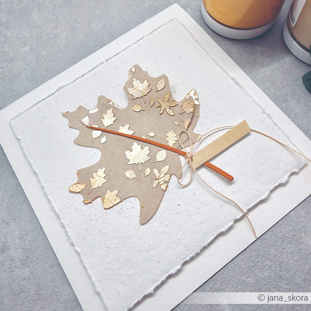 Eine handgefertigte Karte mit einem goldverzierten Papierblatt und einer gebundenen Schnur auf strukturiertem weißem Papier, hergestellt mit der wiederverwendbaren Schablone „Blätterliebe“ – ideal für einzigartige Papierprojekte mit Schablonen.