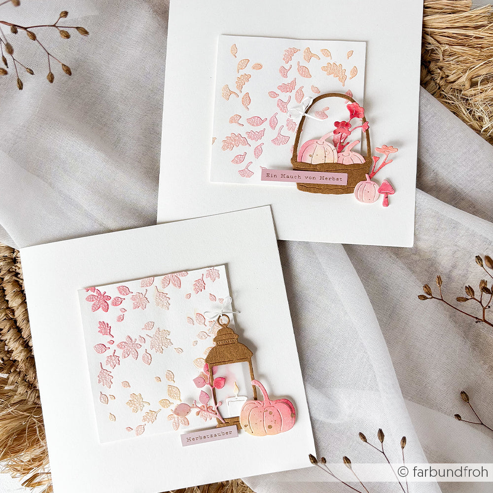 Zwei quadratische Karten mit Herbstmotiven – darunter rosa und goldene Blätter, Kürbisse und Körbe – liegen auf einem weißen Stoffhintergrund. Diese Motive eignen sich ideal für Schablonen-Papierprojekte und fangen die Essenz des Herbstes ein – mit der wiederverwendbaren Schablone „Blätterliebe“ als Inspiration für kreative Bastelprojekte.