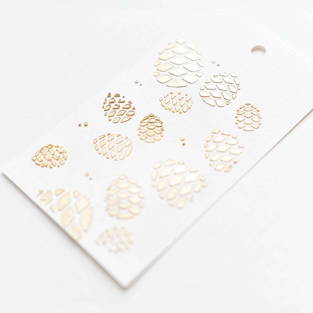 Weißer Anhänger von Alexandra Renke mit Tannenzapfenmotiven aus Goldfolie, inspiriert von Stencil Viele Zapfen, mit einem Loch zum Aufhängen.