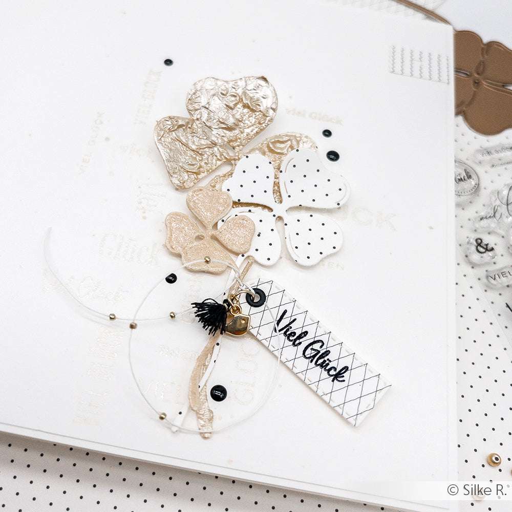 Handgemachte Karte von Alexandra Renke mit strukturierten Kleeblättern aus der Motivstanze Kleeblätter, schwarzen Punkten, einer Quaste und einem "Viel Glück"-Anhänger.