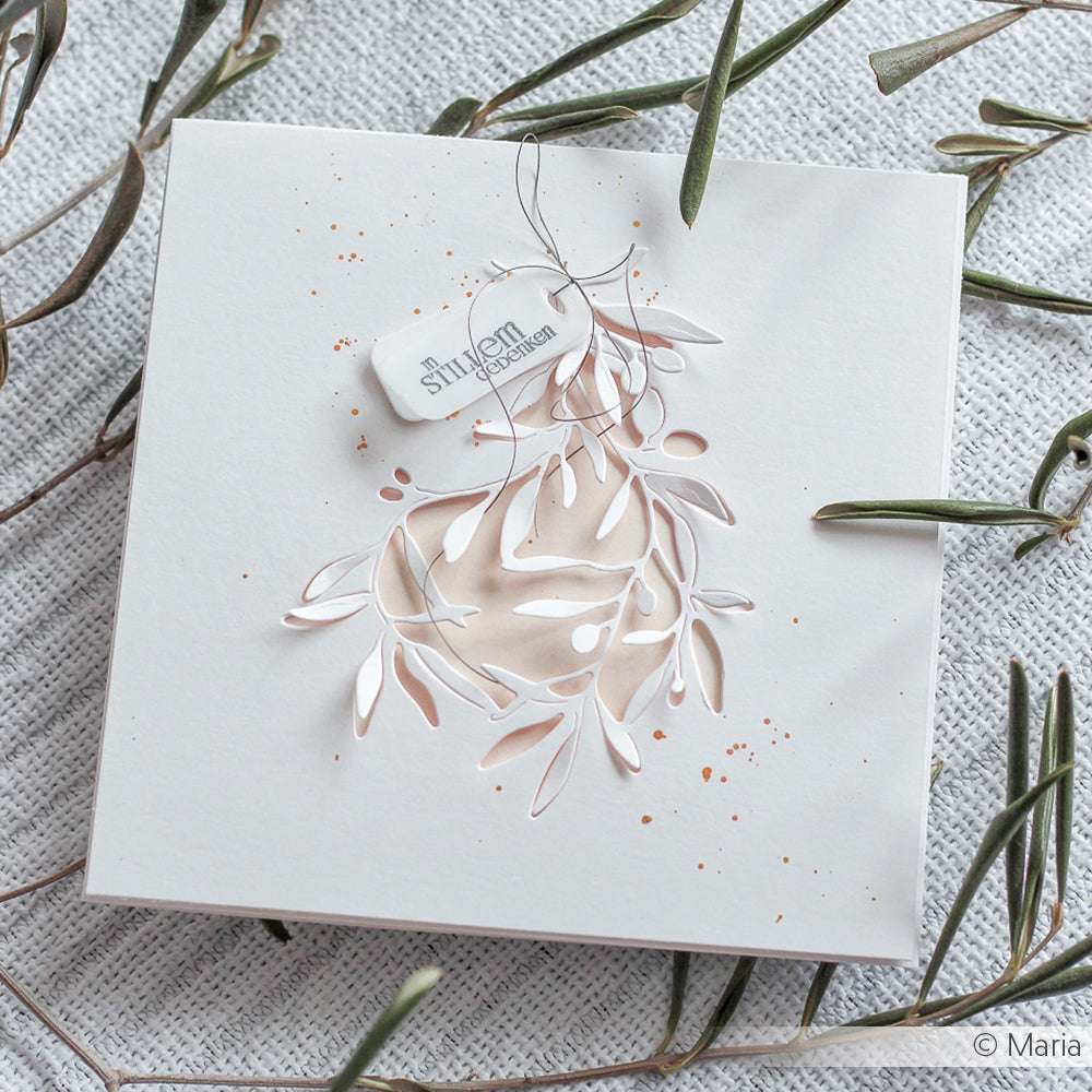 Weiße Karte aus Alexandra Renke-Papier mit zarten, ausgestanzten Blättern, einer Stanze mit Olivenzweigmotiv, einem kleinen Anhänger und kleinen braunen Farbspritzern.