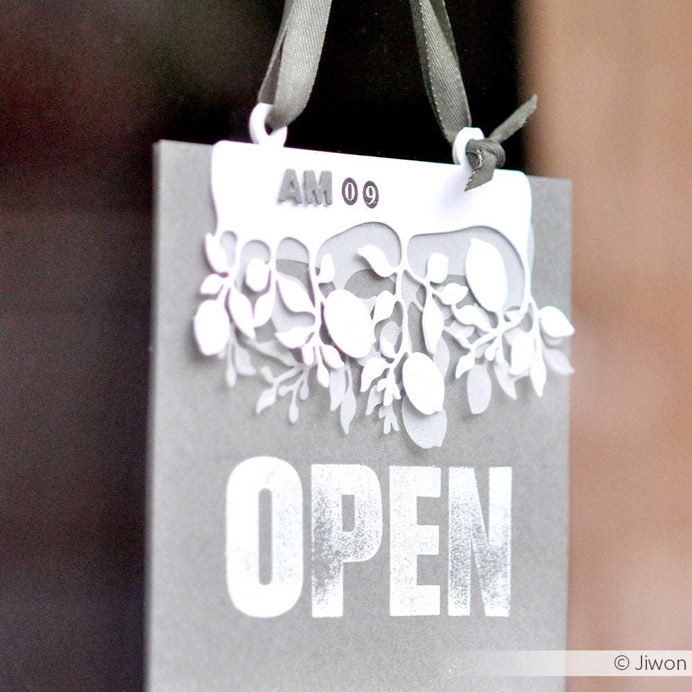 Ein graues OPEN-Schild von Alexandra Renke mit floralen Ausschnitten, die mit komplizierten Stanzformen gefertigt sind, hängt an einem Band; darüber ist AM 09 aufgedruckt.