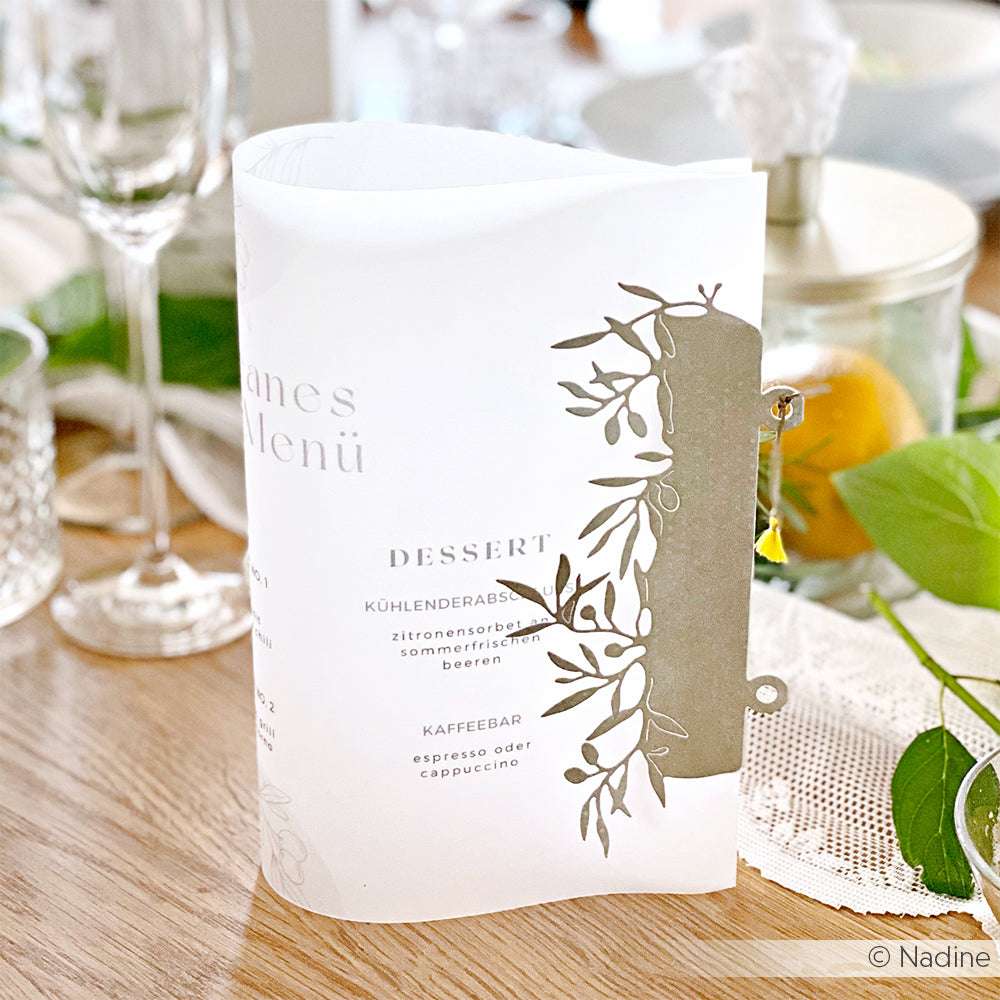 Elegante, weiße, lasergeschnittene Hochzeitsmenükarte von Alexandra Renke mit Blumenmuster, die auf einem dekorierten Esstisch präsentiert wird.