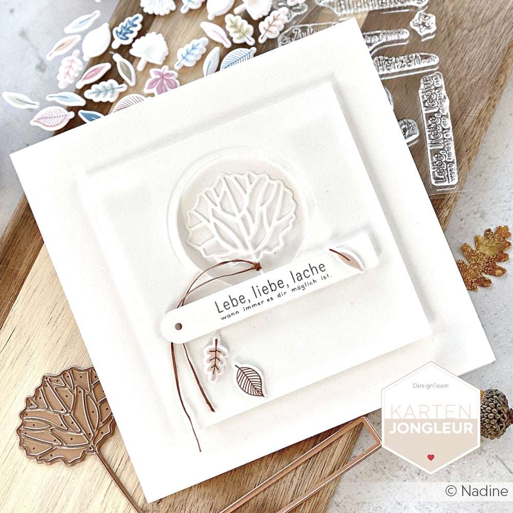 Handgefertigte weiße Karte von Alexandra Renke mit Blattmotiven, die mit DieCut Systemen und Motivstanze hergestellt wurden, deutschem Text und braunen Akzenten, dargestellt auf einem hölzernen Hintergrund.