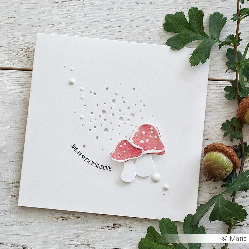 Eine weiße Karte mit zwei roten Pilzen, hergestellt mit der Alexandra Renke Pilz 3 Motivstanze, mit deutschem Text und verziert mit Blättern und Eicheln auf einer Holzoberfläche.