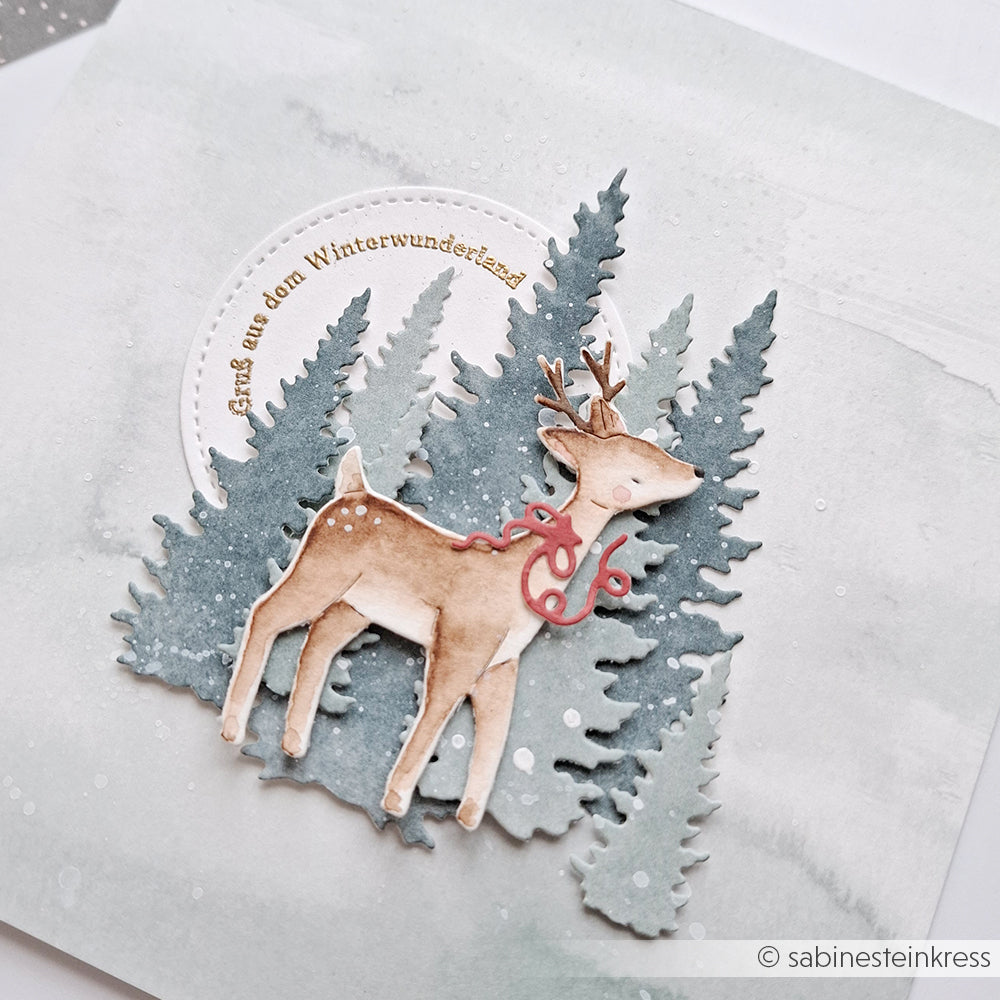 Ein aquarelliertes Rentier mit roter Schleife steht vor verschneiten Immergrünen, die mit der Motivstanze Fünf Tannen von Alexandra Renke auf einer bezaubernden Winterkarte gestaltet wurden.