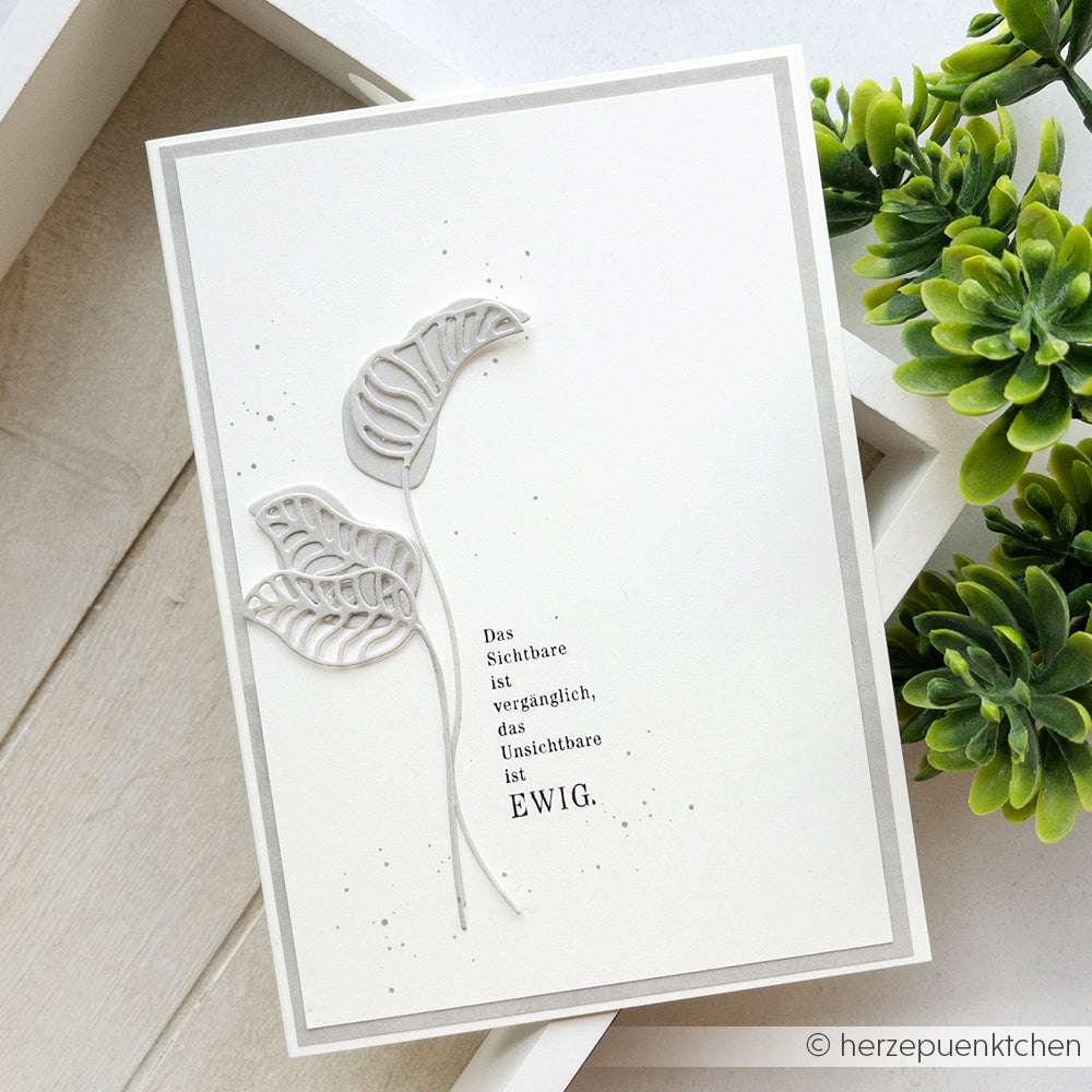 Die minimalistische weiße Karte von Alexandra Renke zeigt zarte Blattausschnitte, die mit einer Motivstanze Blätter erstellt wurden, und ist neben Grünpflanzen auf einer Holzfläche platziert.