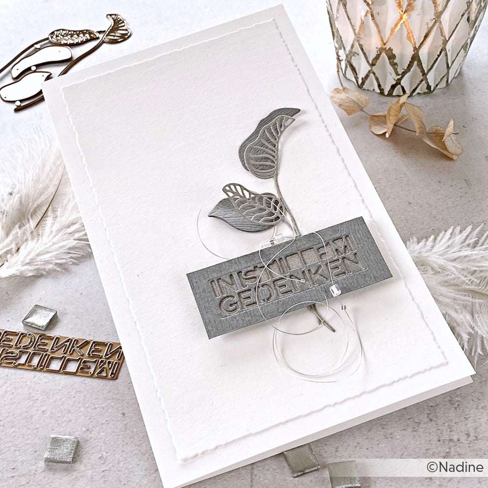 Handgefertigte Karte mit silbernen Blättern, die mit Stanzformen von Alexandra Renke hergestellt wurden, und einer Plakette mit der Aufschrift IN STILLEN GEDENKEN auf weißem, strukturiertem Hintergrund.