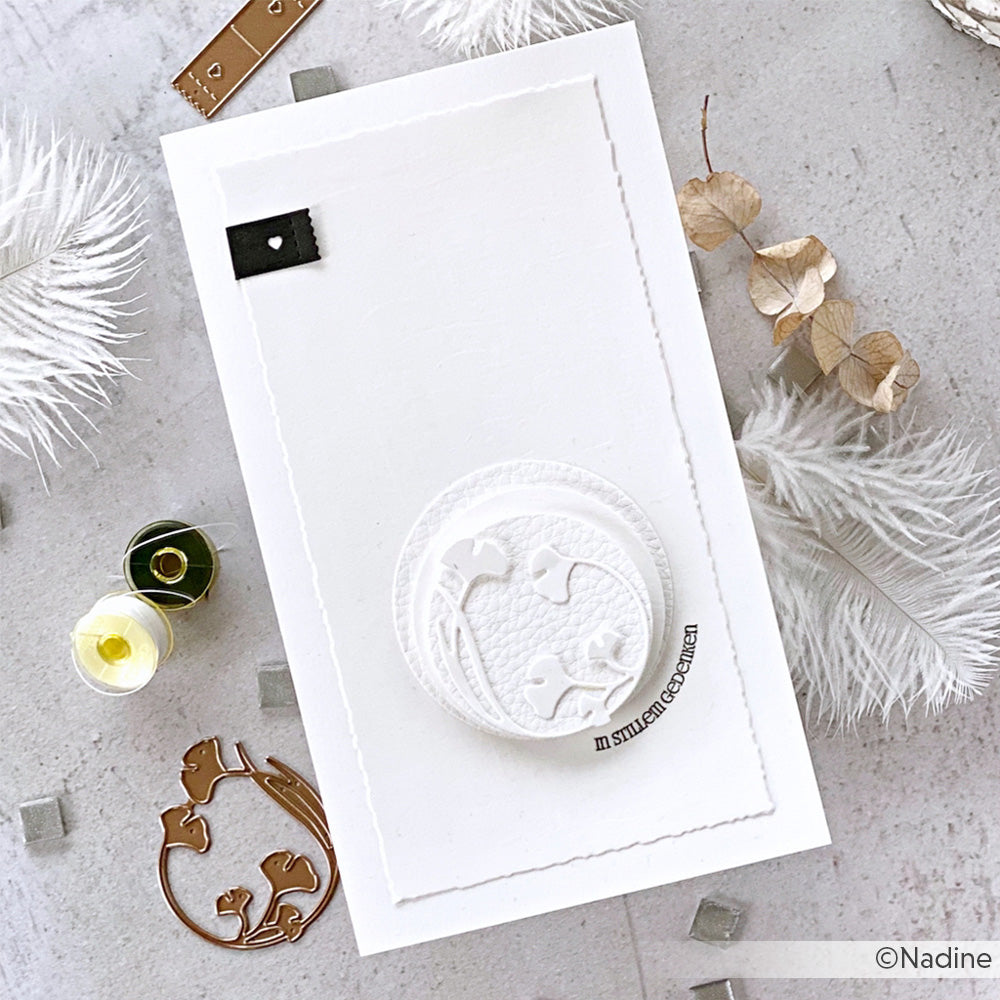 Eine weiße, handgefertigte Karte mit einem erhabenen Blumenkreis, der mit Alexandra Renke Stanzformen hergestellt wurde und mit weißen Federn und getrockneten Blumen verziert ist.