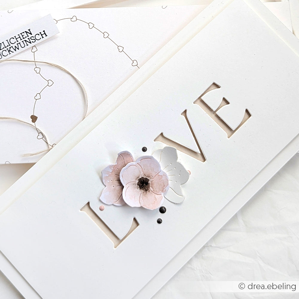 Weiße Karte mit ausgeschnittenem LOVE, verziert mit pastellfarbenen Blumen aus dem Alexandra Renke Motivstanze Quittenblüten Set und Perlen auf Designpapieren, ausgestellt auf einer weißen Fläche.