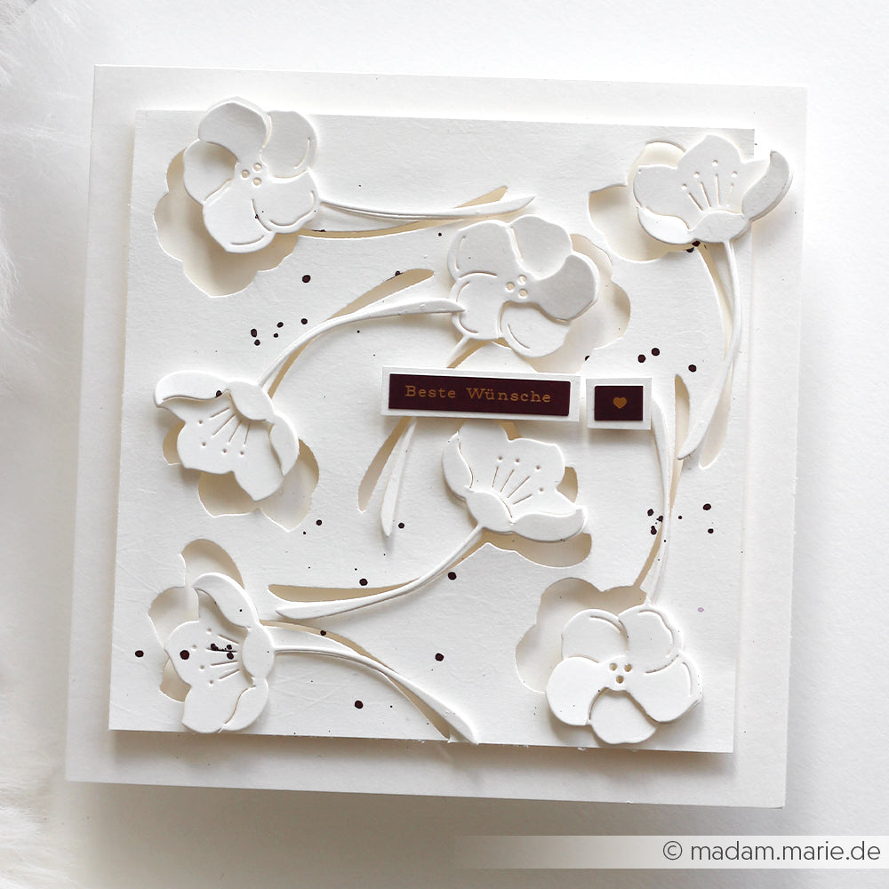Weiße, handgefertigte Karte mit erhabenen Blumenausschnitten aus Alexandra Renke Stanzen für Papier, einem "Beste Wünsche"-Aufkleber und braun gesprenkelten Akzenten.