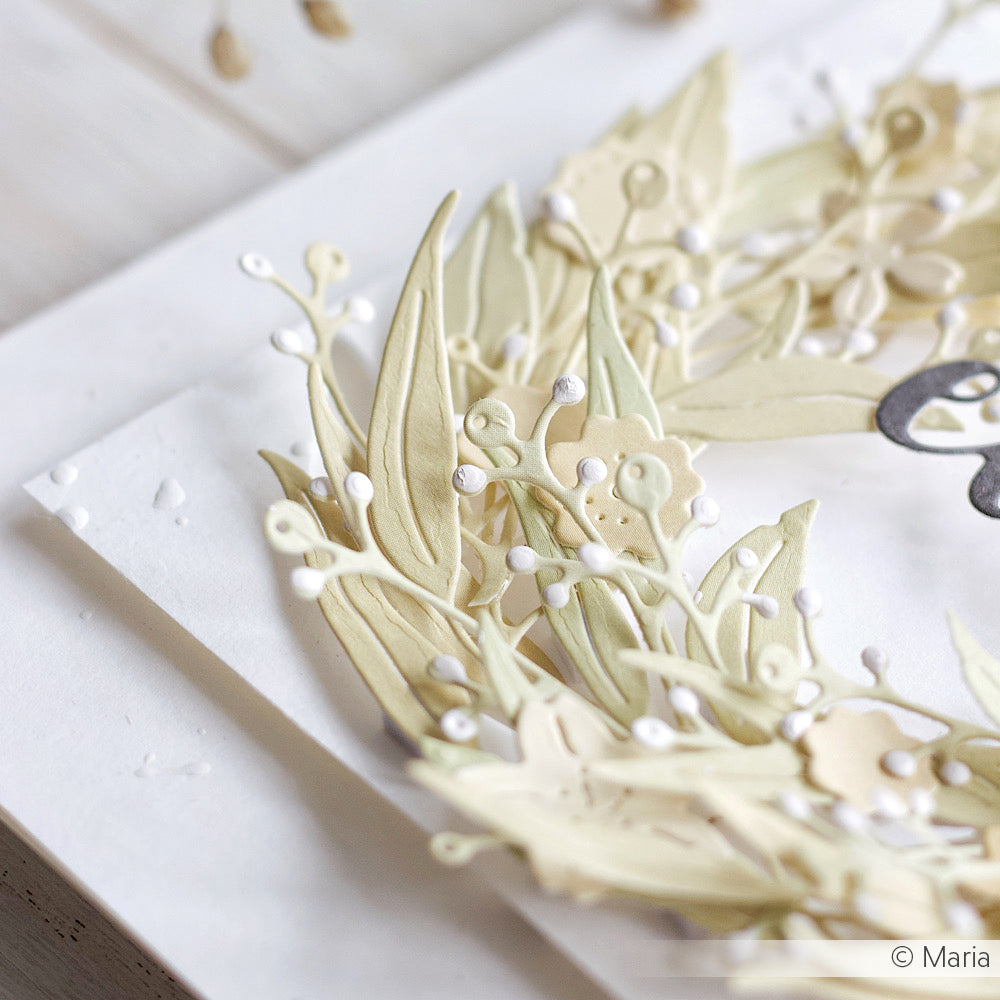 Nahaufnahme eines zarten, geschichteten Papierkranzes, der mit den Werkzeugen von Alexandra Renke hergestellt wurde, mit hellen Blättern und kleinen weißen Beeren auf weißem Hintergrund.