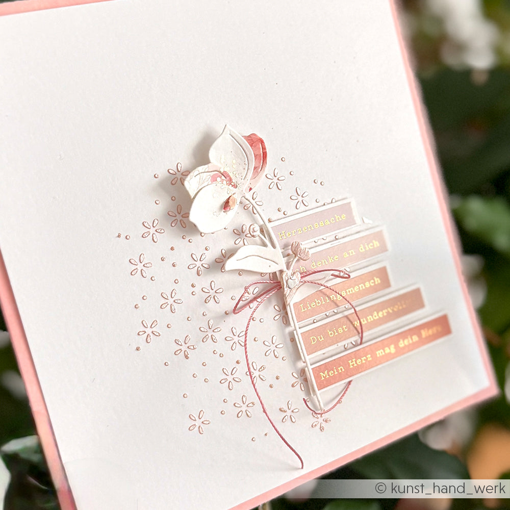 Handgemachte Karte mit einer weißen Blume aus Alexandra Renke Motivstanze Steck-Apfelblüte, rosa Akzenten und deutschen Textstreifen auf elegantem Blumenmusterpapier.