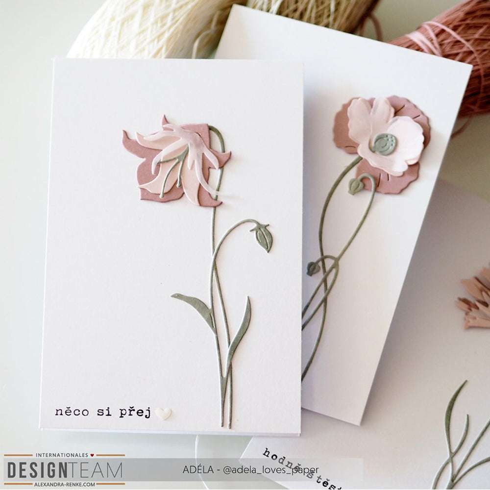 Zwei handgefertigte Grußkarten mit rosafarbenen 3D-Blumen und grünen Stängeln, die mit der Alexandra Renke Schichtblume 11 Motivstanze hergestellt wurden, liegen auf einem Tisch mit Garn im Hintergrund.