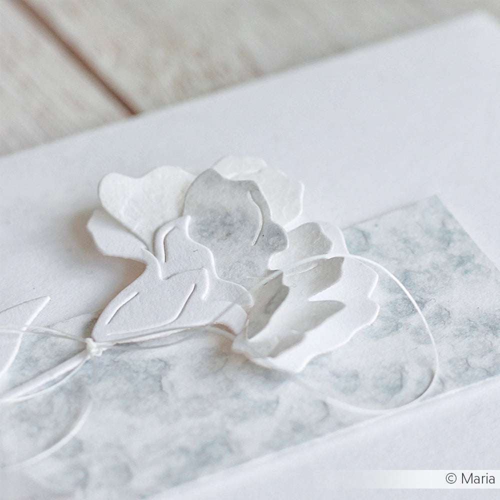 Weiße Papierblumen, die mit der Motivstanze Schichtblume 12 von Alexandra Renke gestaltet wurden, ruhen auf einem strukturierten hellgrauen Hintergrund mit einer dünnen weißen Schnur daneben.