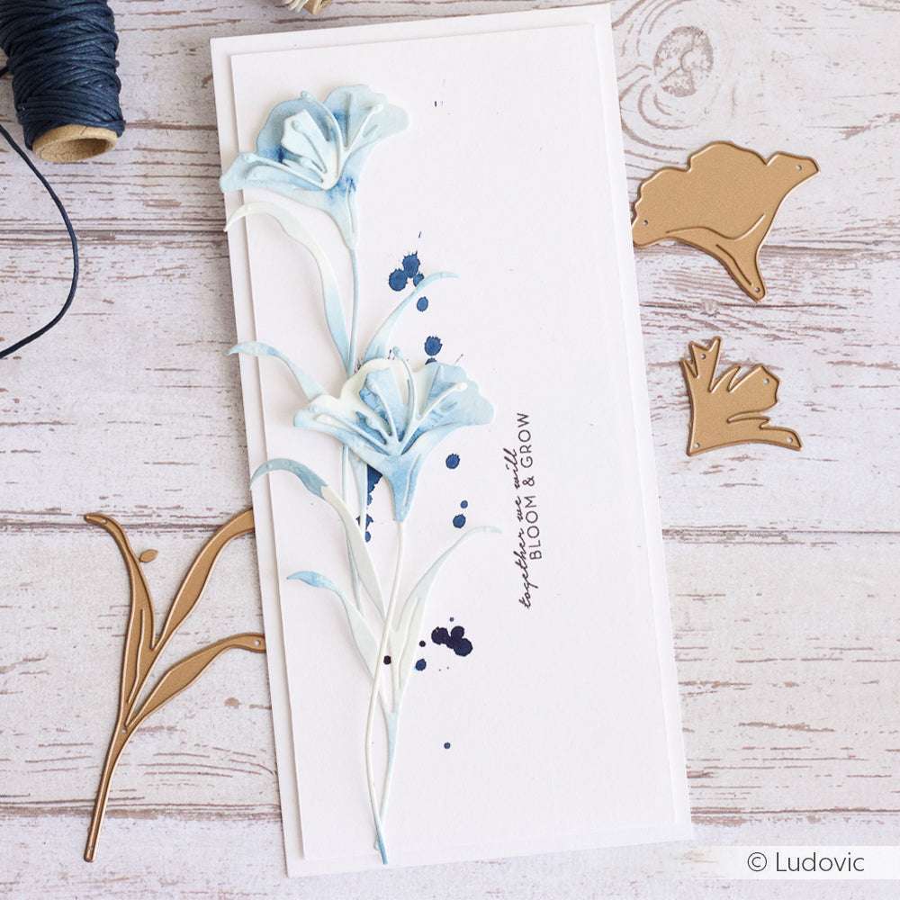 Handgefertigte Karte mit zwei blauen Blumen, Tintenspritzern und goldenen Blumenstanzungen, die mit Alexandra Renke Stanzformen erstellt wurden, dargestellt auf einer Holzoberfläche.