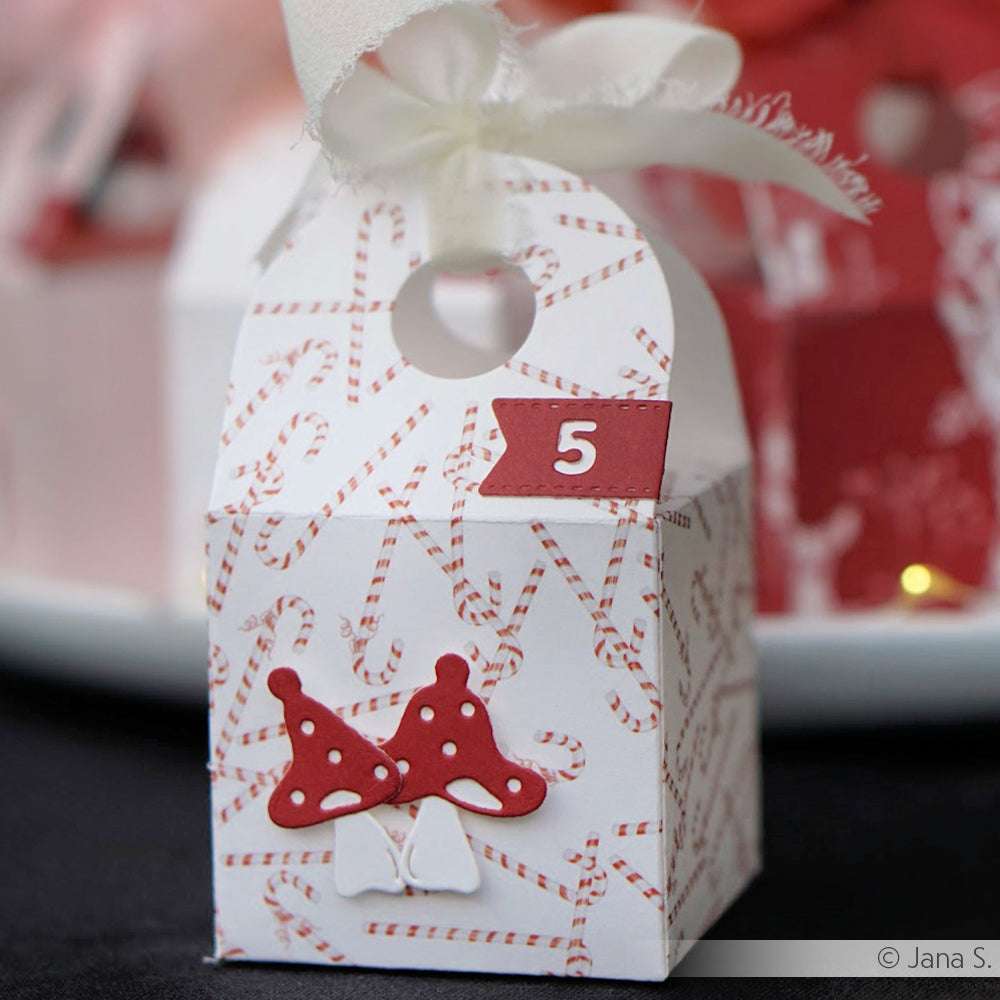 Eine festliche Alexandra Renke Geschenkbox aus Designpapier mit roten Pilzen und Zuckerstangen, veredelt mit weißer Schleife und "5"-Anhänger - ideal für Leckereien, die Sie mit Ihrer Lieblingsmotivstanze Wichtelpilze gebastelt haben.