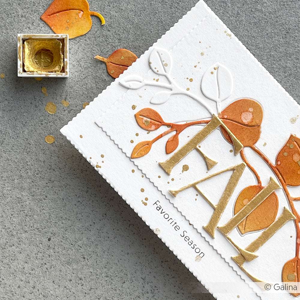 Eine handgefertigte Karte mit Produkten von Alexandra Renke mit goldenen Buchstaben FALL, orangefarbenen Blättern und goldenen Farbspritzern, die mit einer Motivstanze auf einer grauen Oberfläche angebracht wurden.