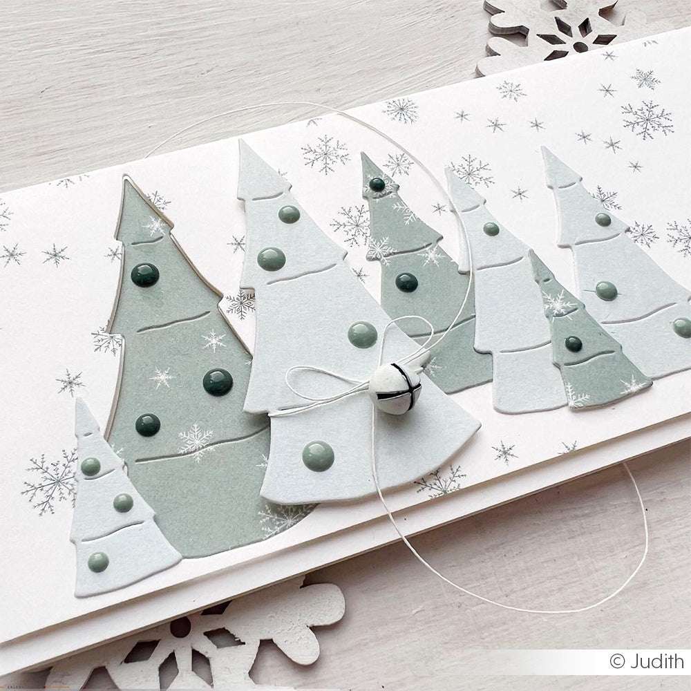 Handgemachte Karte mit geschichteten ausgeschnittenen Bäumen unter Verwendung der Alexandra Renke Motivstanze Kleiner Tannenwald 2, Schneeflocken, grünen Punkten auf Premium Designpapieren, sowie einer weißen Schnur und einem Glöckchen.