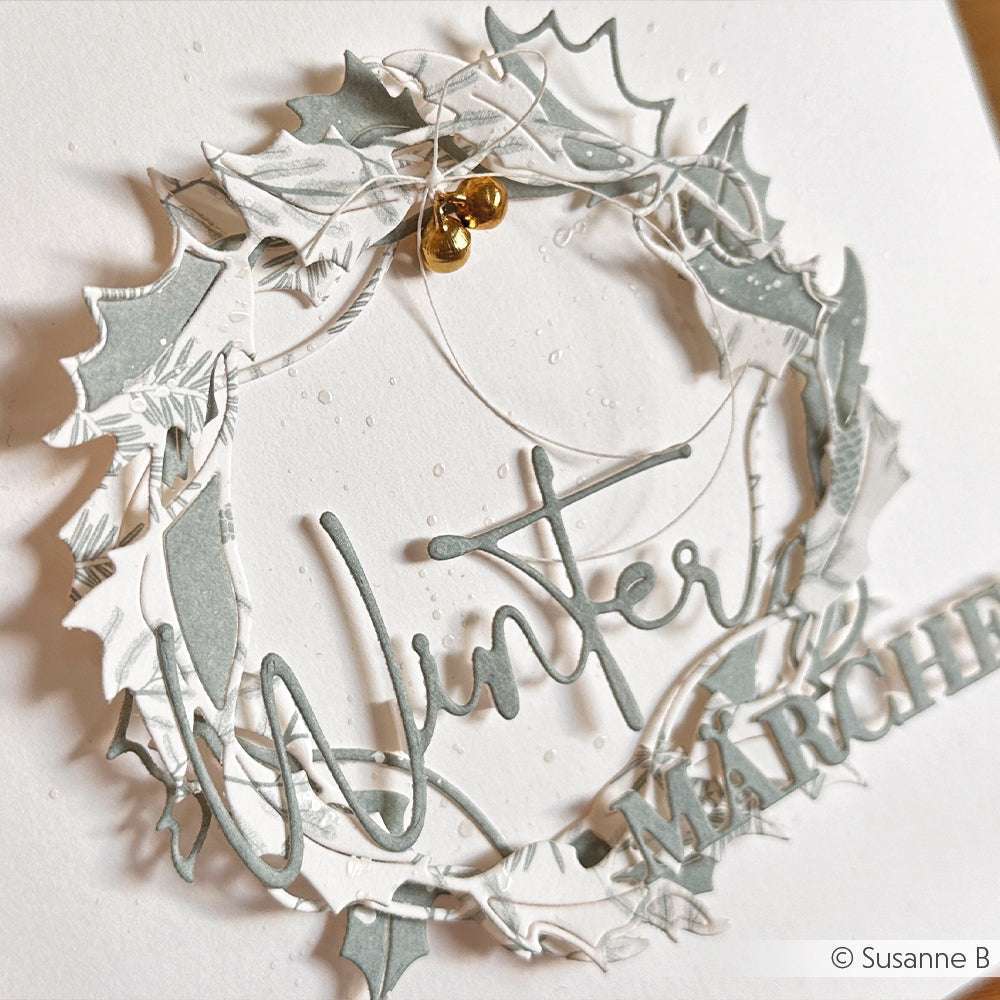 Handgemachte Karte von Alexandra Renke mit einem Papierkranz, der mit Designpapieren und Motivstanze Ilexkranz DieCut Systeme hergestellt wurde, goldenen Glocken und dem Text "Winter Marché" auf weißem Hintergrund.
