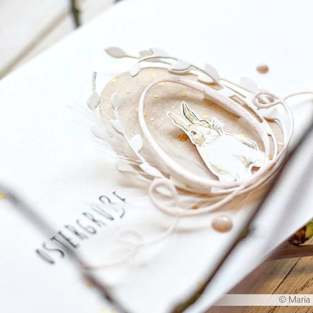 Handgefertigte Osterkarte von Alexandra Renke mit einer Hasenillustration, zarten Blättern aus Stanzsystemen und Motivstanze Weidenkätzchenbögen und dem Text "OSTERGRÜßE" auf weichen, neutralen Designpapieren.