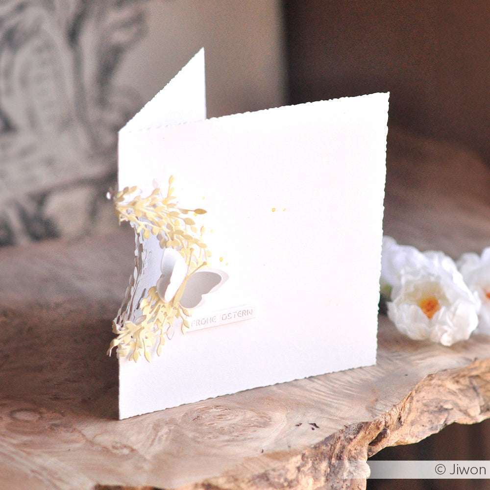 Weiße handgefertigte Karte von Alexandra Renke mit einem Schmetterlings- und Blumenmotiv, hergestellt mit Designpapieren und einer Motivstanze Weidenkätzchenkranz, auf einer rustikalen Holzoberfläche.