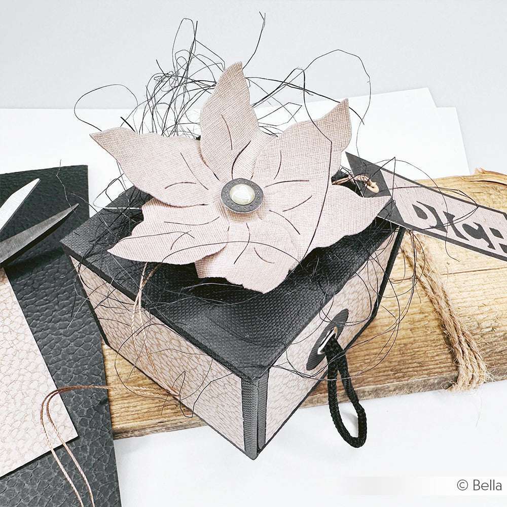 Eine schwarze Geschenkschachtel mit einer strukturierten Blume, die mit der Motivstanze Blumen-Windrad von Alexandra Renke gefertigt wurde, sitzt elegant auf einer rustikalen Holzoberfläche.
