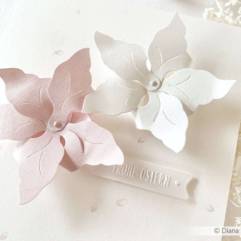 Zwei pastellfarbene Papierblumen, gestanzt mit der Blumen-Windrad-Stanze von Alexandra Renke und mit Perlen in der Mitte, zieren eine weiße Karte mit der Aufschrift "Frohe Ostern". Perfekt für Ostergrüße, glänzen diese Blüten auf hochwertigem Designpapier.