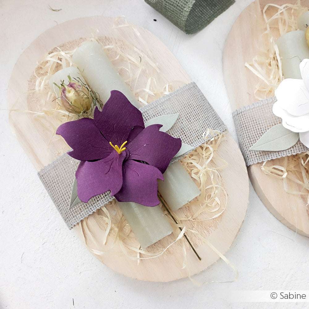 Eine gerollte Kerze mit einer violetten Papierblume von Alexandra Renke Designpapiere und getrockneten Rosen, eingewickelt in Sackleinen, ruht auf einem Holztablett mit Stroh.