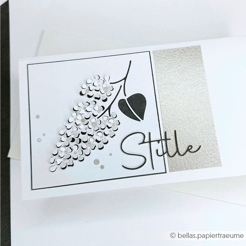 Weiße Karte mit silbernem Blumendesign, hergestellt mit Alexandra Renke Motivstanze Negativ-Blume 2 und Designpapieren, mit dem Wort "Stille" in eleganter Schrift und einer strukturierten Silberplatte.