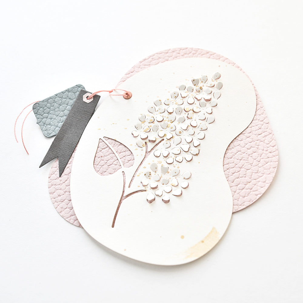 Eine zarte Blumenkarte von Alexandra Renke mit ausgeschnittenen Blumen aus Stanzformen, verziert mit Bandanhängern und pastellfarbenen, strukturierten Formen auf weißem Hintergrund.