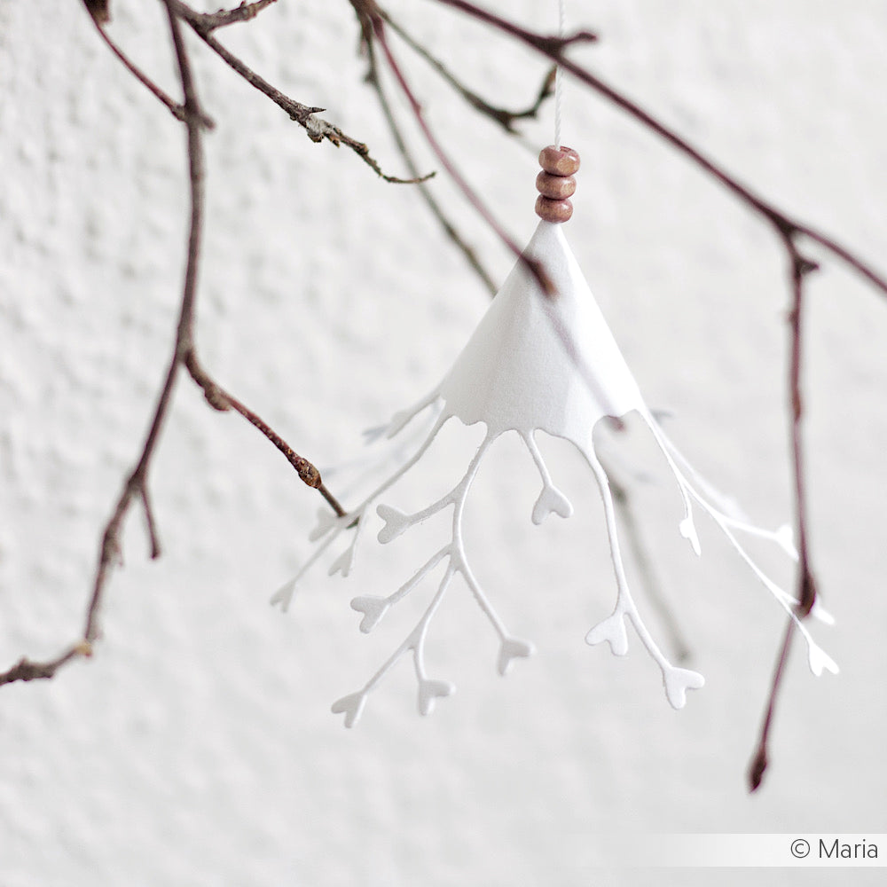 Das weiße Papierornament von Alexandra Renke, das mit Stanzsystemen und der Motivstanze Herzchen-Anhänger hergestellt wurde, hat zarte astähnliche Ausschnitte und hängt elegant an einem kahlen Zweig vor einem hellen Hintergrund.