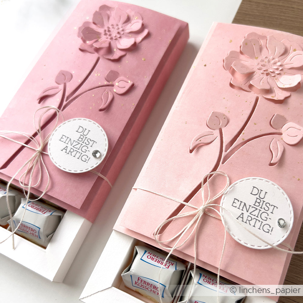 Zwei rosafarbene Geschenkschachteln mit Blumenprägung von Alexandra Renke, die mit Pralinen gefüllt und mit einer Schnur und einem "DU BIST EINZIGARTIG!"-Anhänger verschnürt sind, werden aus elegantem Designpapier hergestellt und erhalten so eine ganz besondere Note.