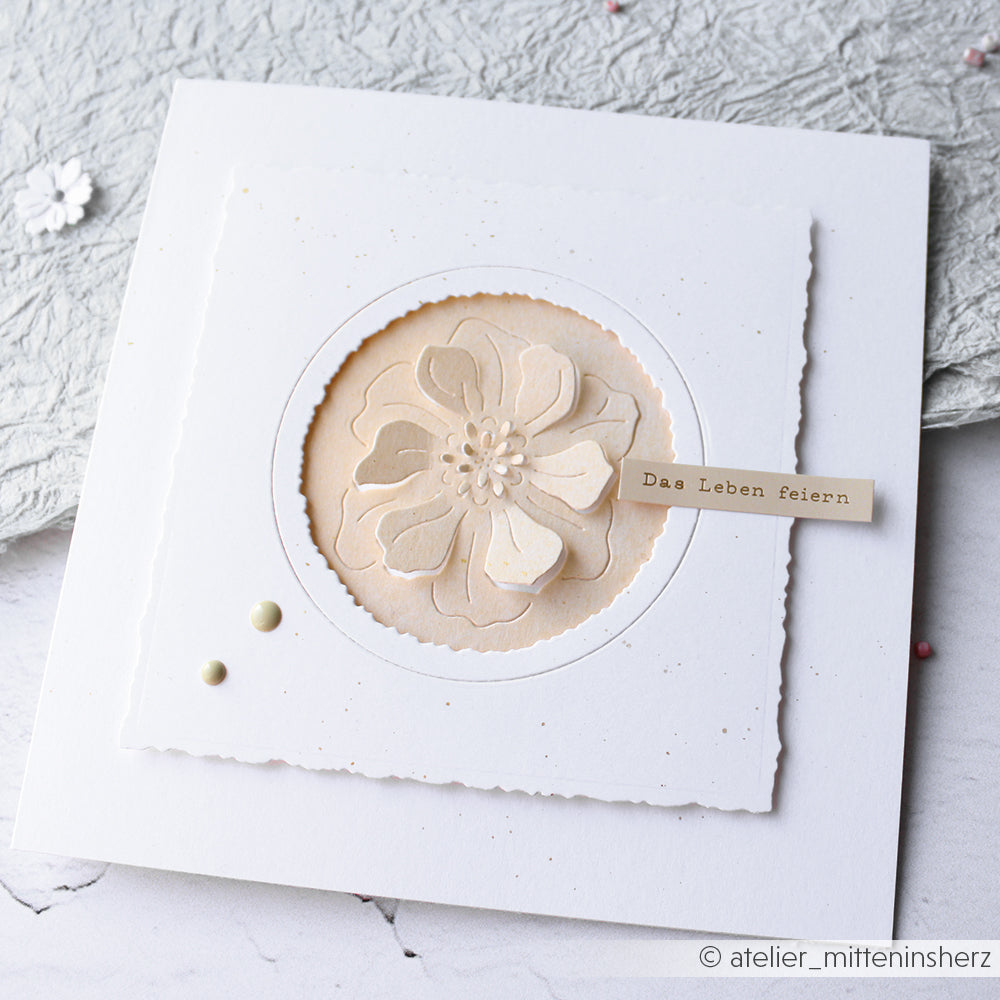 Handgefertigte weiße Karte von Alexandra Renke, hergestellt mit Designpapier und einer negativen Blumenstanze. Mit einer geschichteten beigen Blume und einem Schildchen mit der Aufschrift "Das Leben feiern" auf Deutsch.