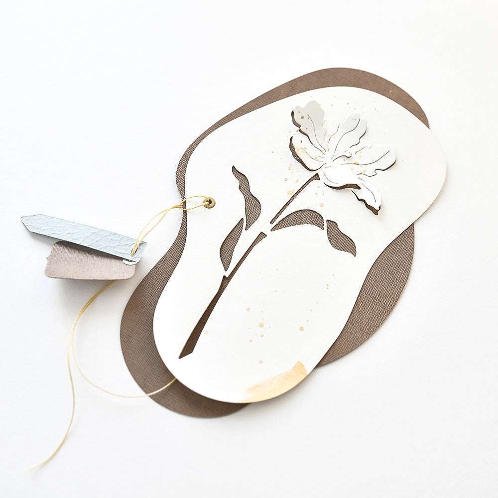 Ein Lagenpapier-Anhänger von Alexandra Renke mit einem Negativ-Blume 5-Blumenausschnitt, braunem Hintergrund und zwei kleinen Anhängern, die mit Schnur gebunden sind.