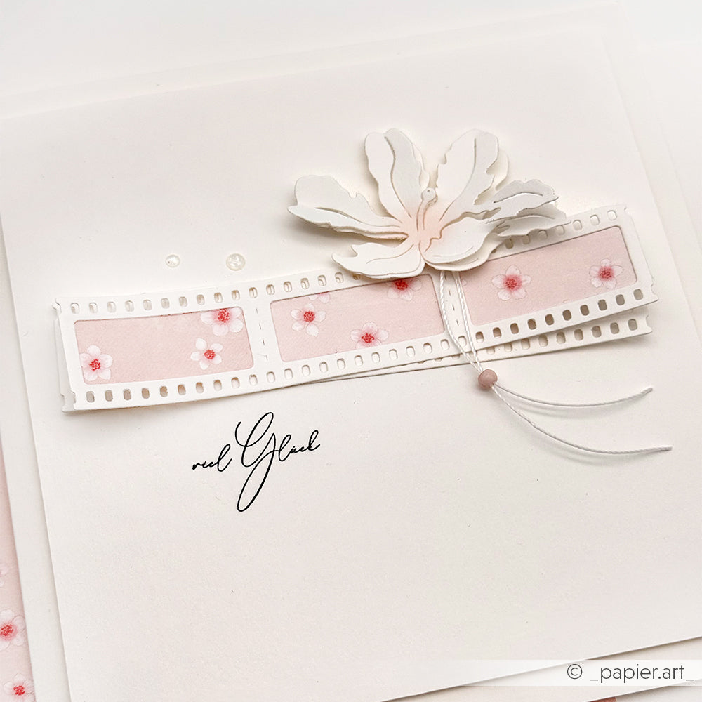 Handgefertigte Karte mit weißer 3D-Blüte und rosafarbenem Folienstreifen, hergestellt mit Alexandra Renke DieCut Systeme Motivstanze Negativ-Blume 5, akzentuiert durch kleine rosa Blüten, zarte Fäden und elegantes Designpapier.
